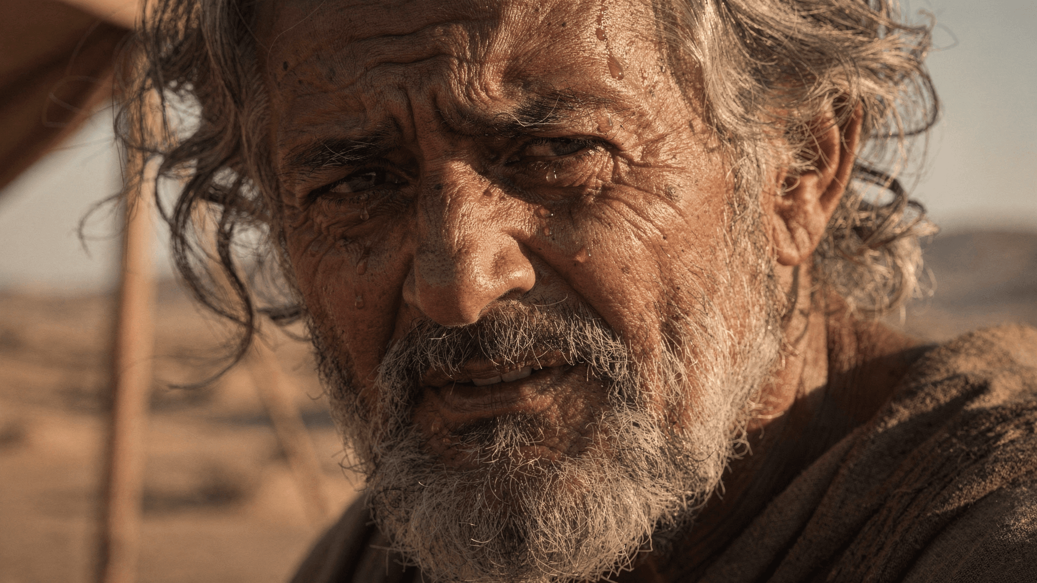 This video features a close-up, static shot of an older man with a weathered face, graying beard, and curly hair. The overall mood is somber and reflective, with a dramatic emotional tone. The setting appears to be an outdoor, arid environment, possibly a desert, indicated by the sandy background and harsh lighting. The visual style is realistic and cinematic, emphasizing the man's detailed facial features, including wrinkles and sweat beads. The color palette consists of warm, earthy tones like browns, ochres, and muted yellows, with high saturation. The strong lighting creates deep shadows and highlights, enhancing the texture of his skin and hair.